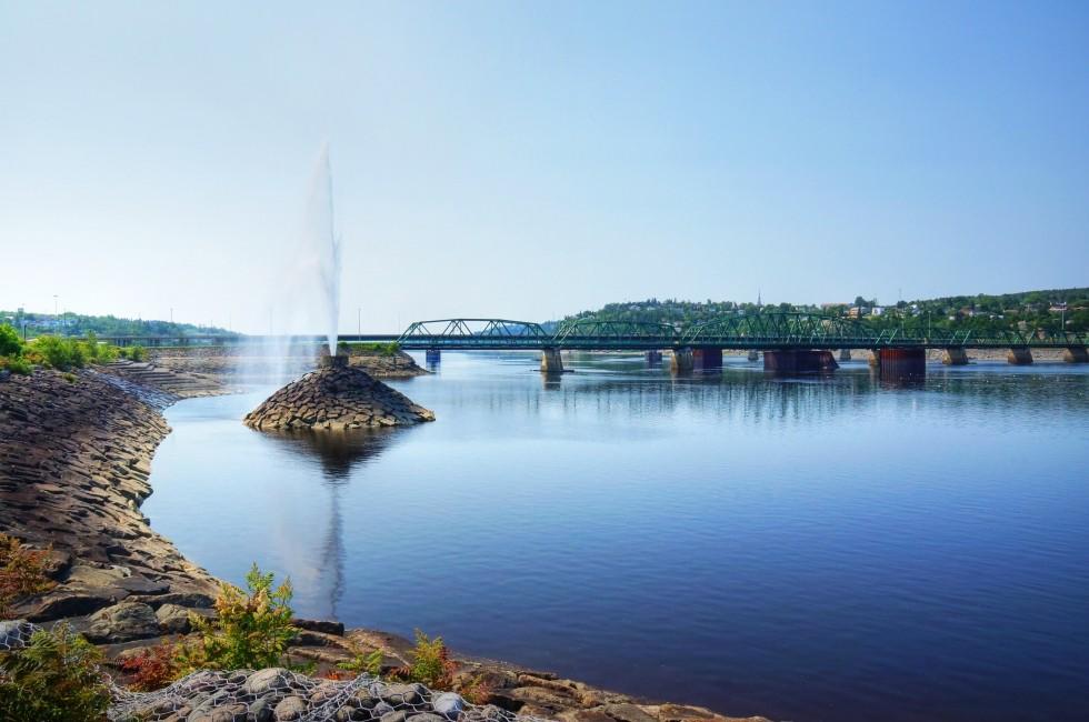 Beautiful water fountain in a river by a bridge, public park in Saguenay, Quebec, Canada; 