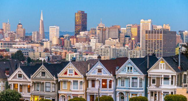 The Painted Ladies of San Francisco, California sit glowing amid the backdrop of a sunset and skyscrapers.