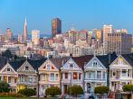 The Painted Ladies of San Francisco, California sit glowing amid the backdrop of a sunset and skyscrapers.