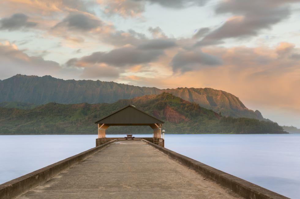 Rising sun illuminates the peaks of Na Pali mountains over the calm bay and Hanalei Pier in long exposure photo.