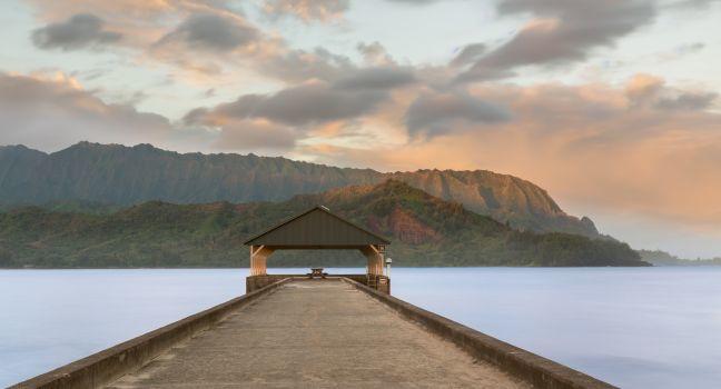 Rising sun illuminates the peaks of Na Pali mountains over the calm bay and Hanalei Pier in long exposure photo.